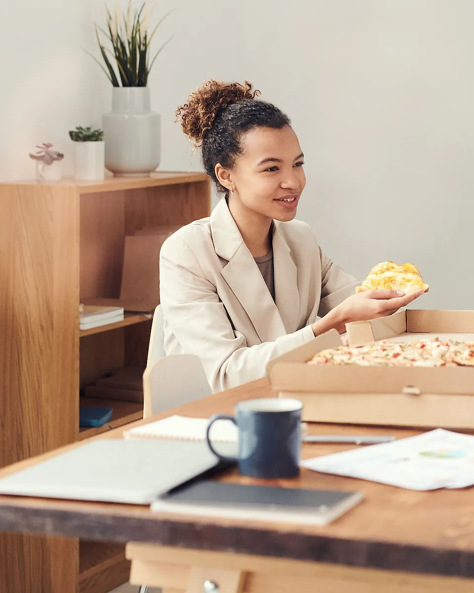 En kvinne i blazer holder et pizzastykke ved et bord på kontoret