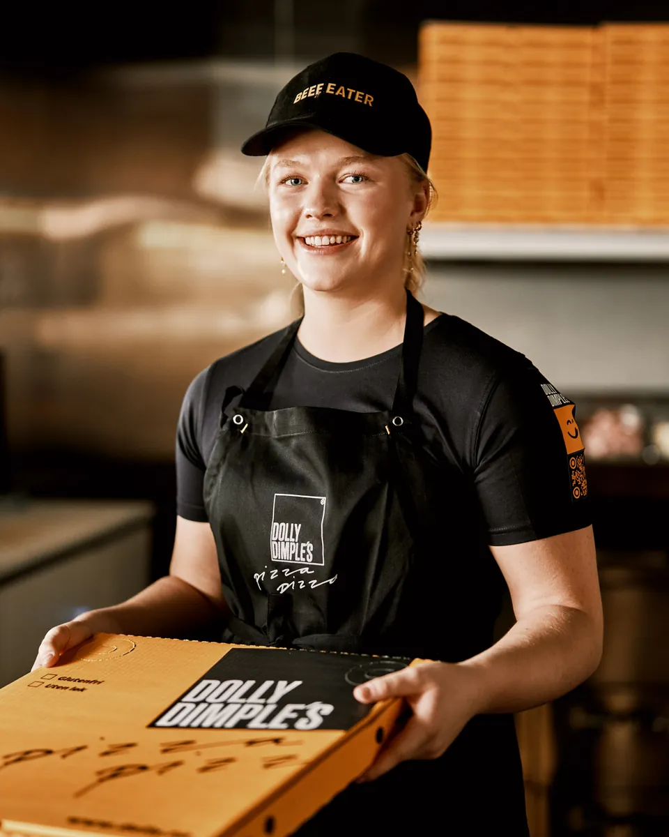 En smilende ansatt hos Dolly Dimples holder en pizzaeske med Dolly-logoen